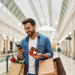 A man holding Valentine's chocolates and paper shopping bags with red flowers in one while smiling at his phone